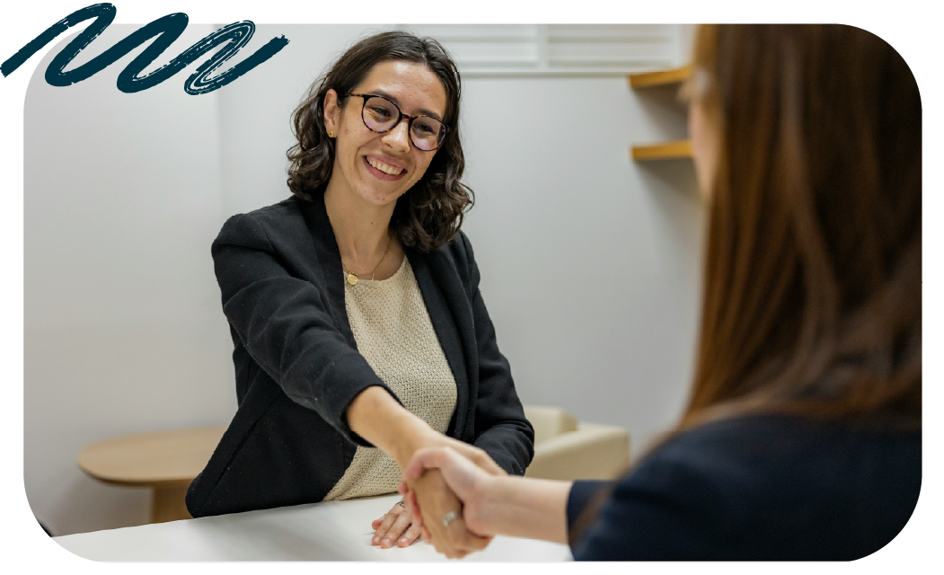 A professional in glasses and a blazer shaking hands with someone across a desk in an office.