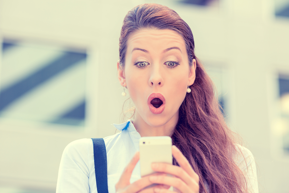 Closeup portrait anxious young girl looking at phone seeing bad news or photos with disgusting emotion on her face isolated outside city background. Human emotion, reaction, expression